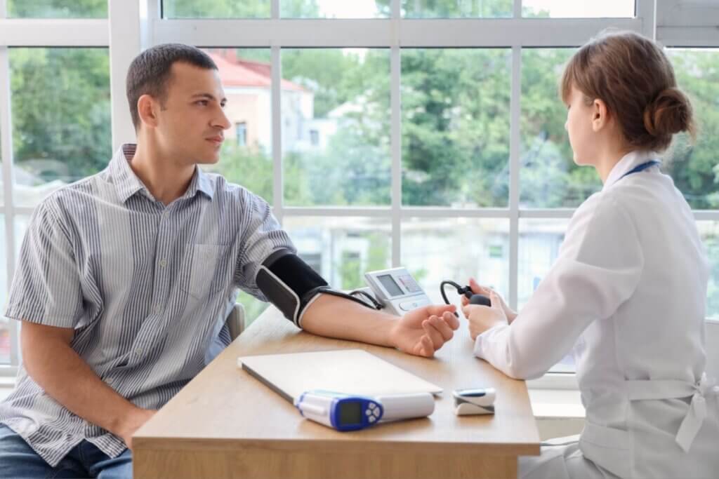 Homem com cabelo curto, usando camisa cinza listrada medindo a pressão com acompanhamento de médica usando coque e jaleco branco em consultório