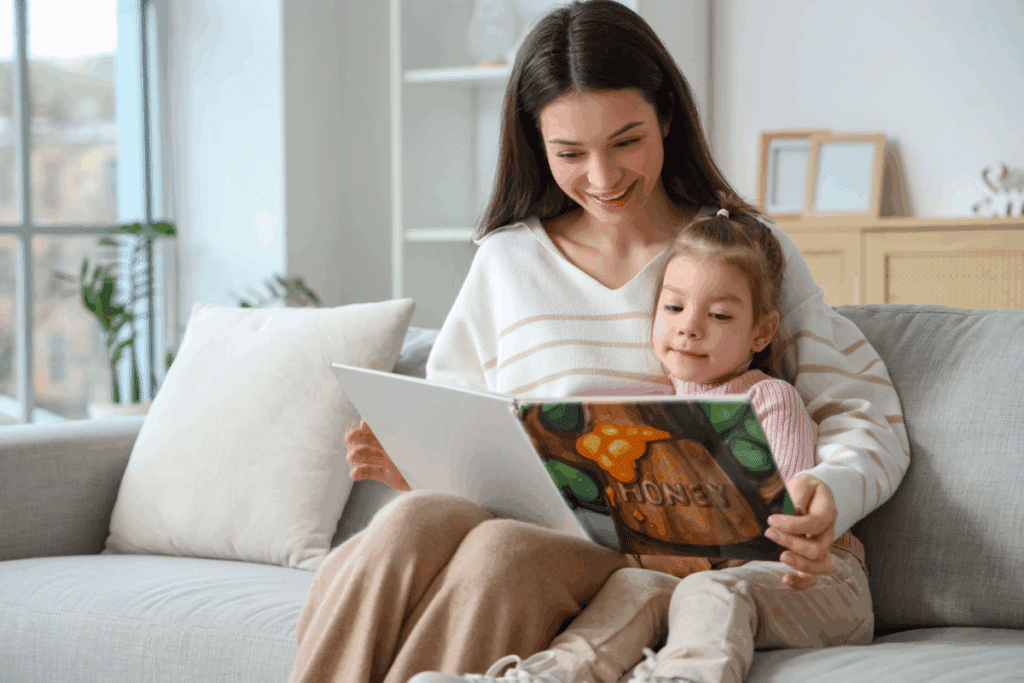 Mulher com cabelo liso, longo, solto e usando camisa branca com listras em bege e calça bege, lendo livro ilustrado para menina com cabelo preso, usando camiseta rosa e calça bege