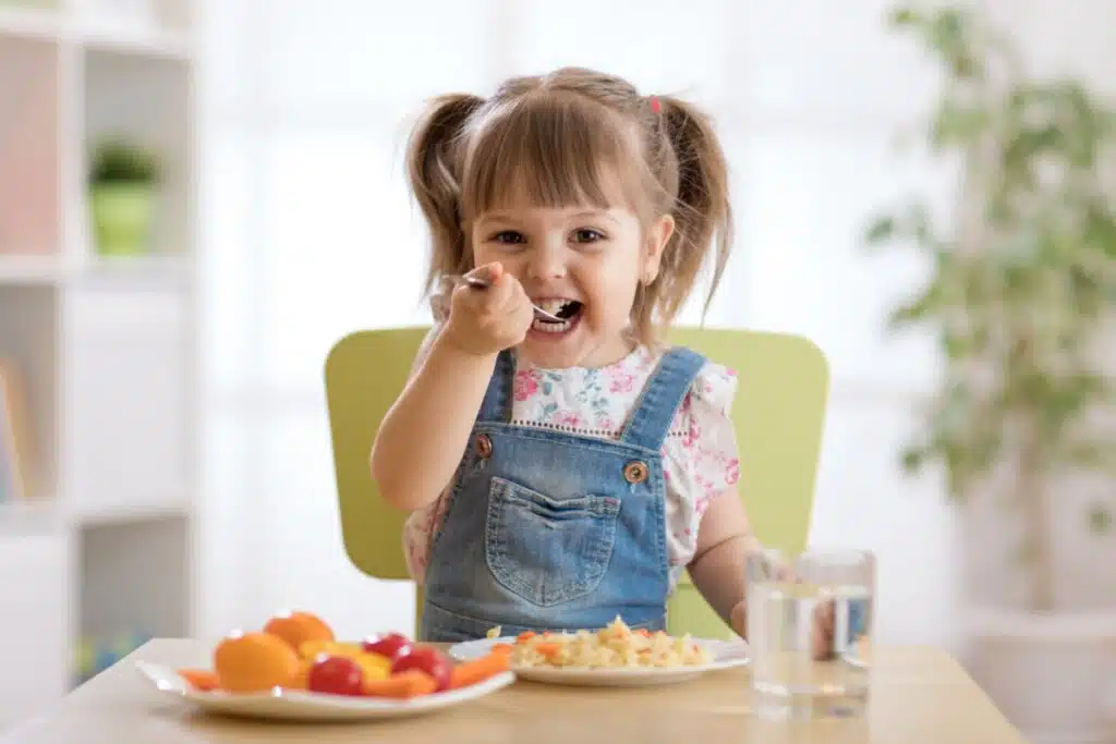 menina pequena vestindo jardineira azul e levando talher com comida à boca. ela está sentada à mesa com prato com comida
