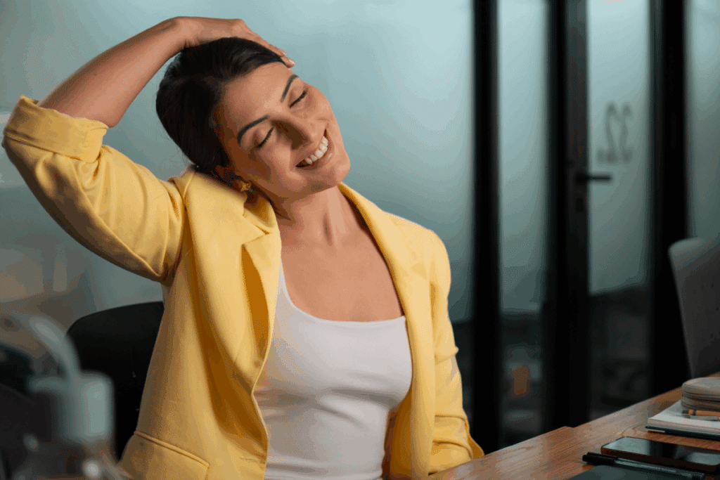 Mulher, usando blusa e blazer amarelo, sentada em um escritório, alongando o pescoço e sorrindo