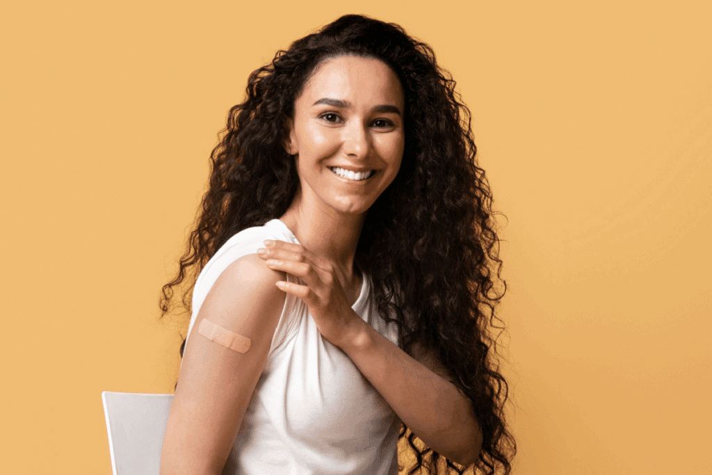 Mulher com cabelo longo, cacheado, usando camiseta branca mostrando curativo após tomar vacina em fundo laranja-claro
