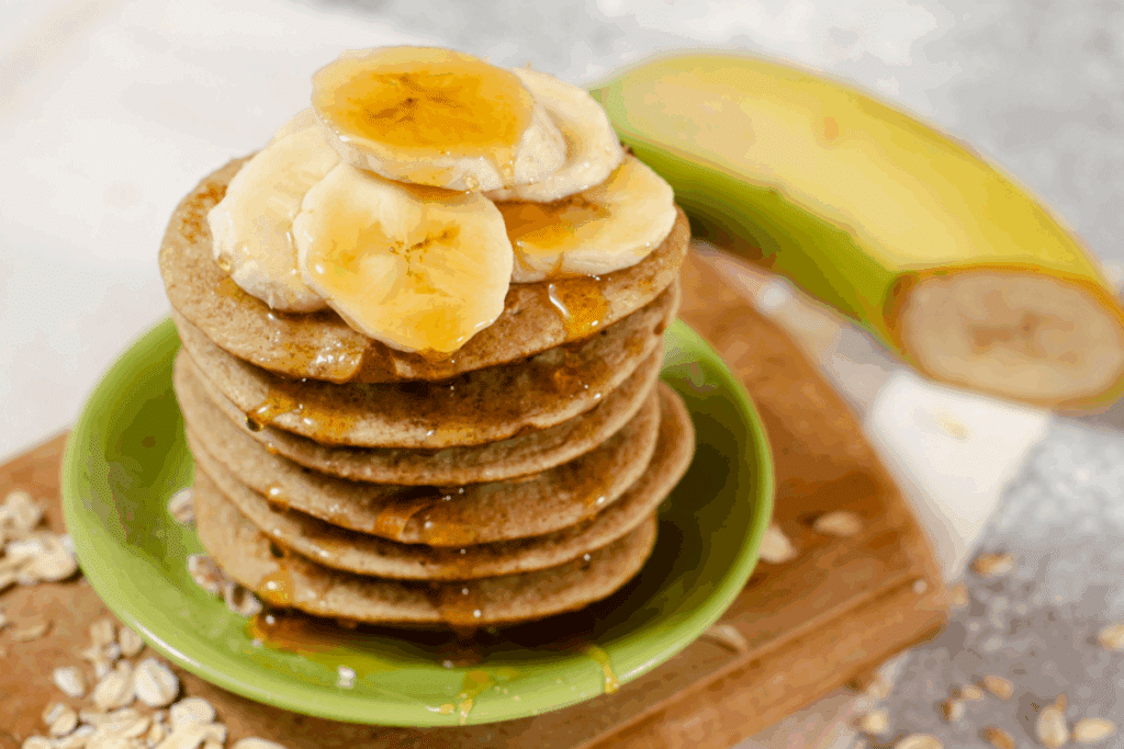 Panqueca de banana com aveia, ovo e mal empilhadas em prato verde em cima de tábua de madeira com rodelas de banana por cima