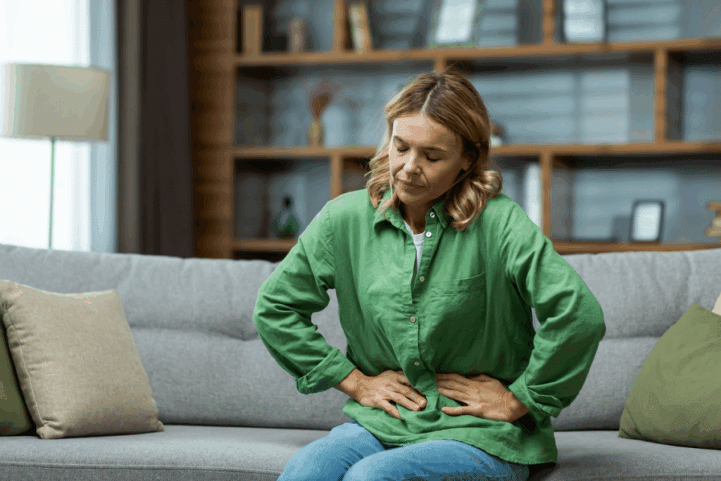 Mulher adulta de cabelos loiros e curtos, vestindo uma camisa verde de manga comprida e calça jeans, sentada no centro de um sofá cinza em uma sala de estar. Ela está levemente curvada para a frente, com os olhos fechados e ambas as mãos pressionadas sobre o abdômen, demonstrando uma expressão de dor ou desconforto abdominal. Ao fundo, vê-se uma estante de madeira desfocada com livros e objetos decorativos.