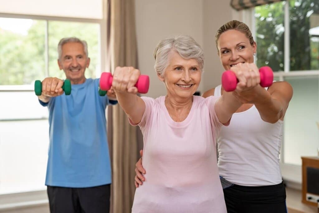 Idosos praticando exercícios de força com halteres coloridos sob orientação de uma profissional de saúde