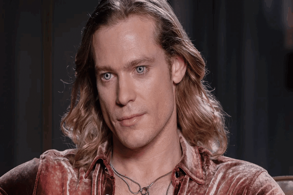 Homem com o cabelo loiro, na altura do ombro, olhos azuis usando camisa de veludo marrom