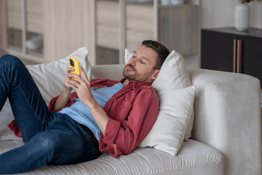 homem de camiseta azul e camisa vermelha, deitado em sofá branco e mexendo em celular amarelo