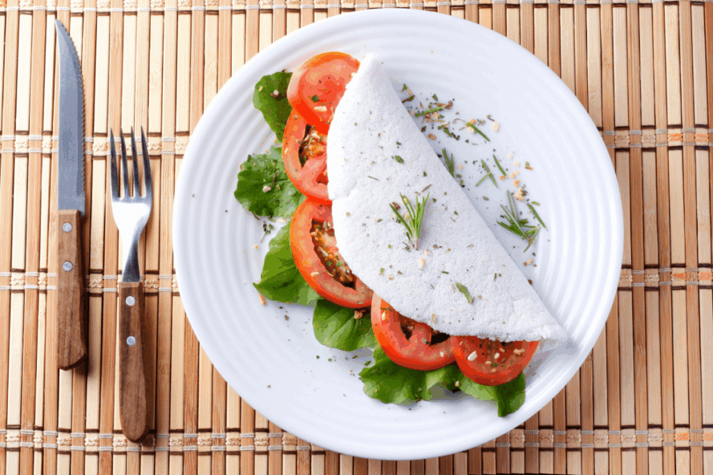 Tapioca em prato branco com recheio de tomate e folhas verdes em cima de jogo americano de bambu ao lado de talheres