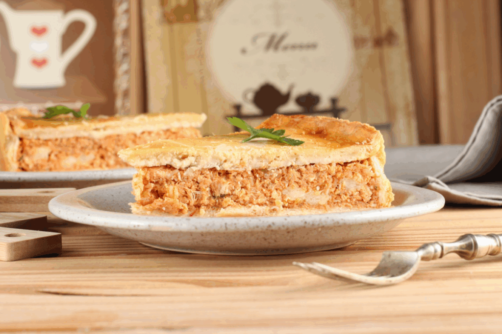 Fatia de torta de frango servida em um prato sobre uma mesa de madeira, com recheio cremoso e bem recheado, envolto por uma massa dourada e crocante. Ao fundo, a torta inteira aparece parcialmente cortada, destacando uma receita caseira