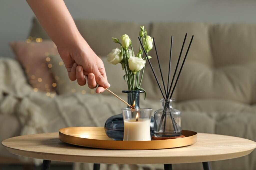 A imagem mostra uma mão acendendo uma vela sobre uma mesa de centro, criando um ambiente acolhedor. Ao lado da vela, há pequenos vasos decorativos com flores brancas e um difusor de aromas com varetas, organizados em uma bandeja. Ao fundo, um sofá desfocado reforça a sensação de conforto e tranquilidade do ambiente doméstico.