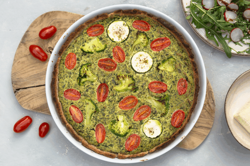 Quiche de espinafre com brócolis e abobrinha servido em refratário branco em cima de tábua de madeira em cima de bancada branca