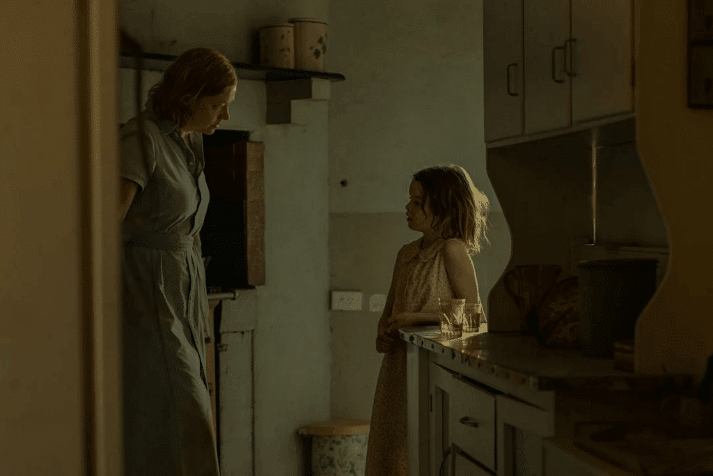 Uma mulher de vestido claro em pé em uma cozinha rústica olha seriamente para uma garotinha em frente ao balcão.
