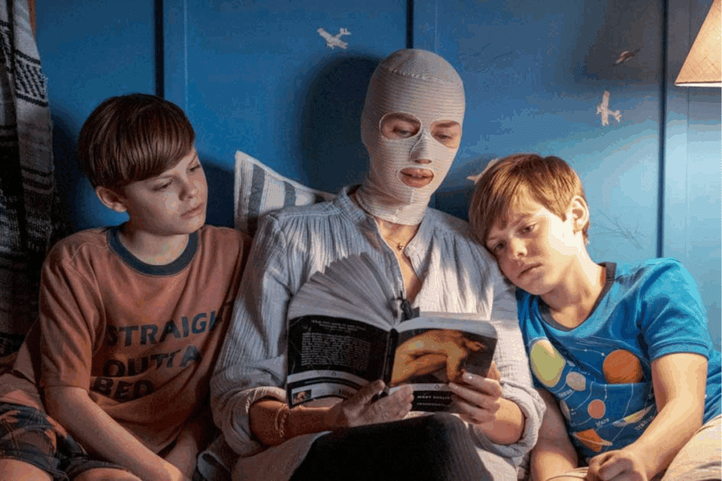 Mulher com faixa no rosto, deitada entre dois meninos enquanto lê um livro para eles 