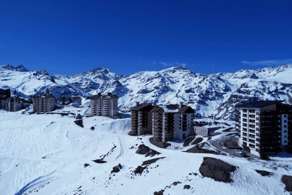 Vista panorâmica de um resort de esqui com vários edifícios e hotéis instalados em encostas cobertas de neve, com as montanhas imponentes e nevadas da Cordilheira dos Andes ao fundo sob um céu azul limpo.