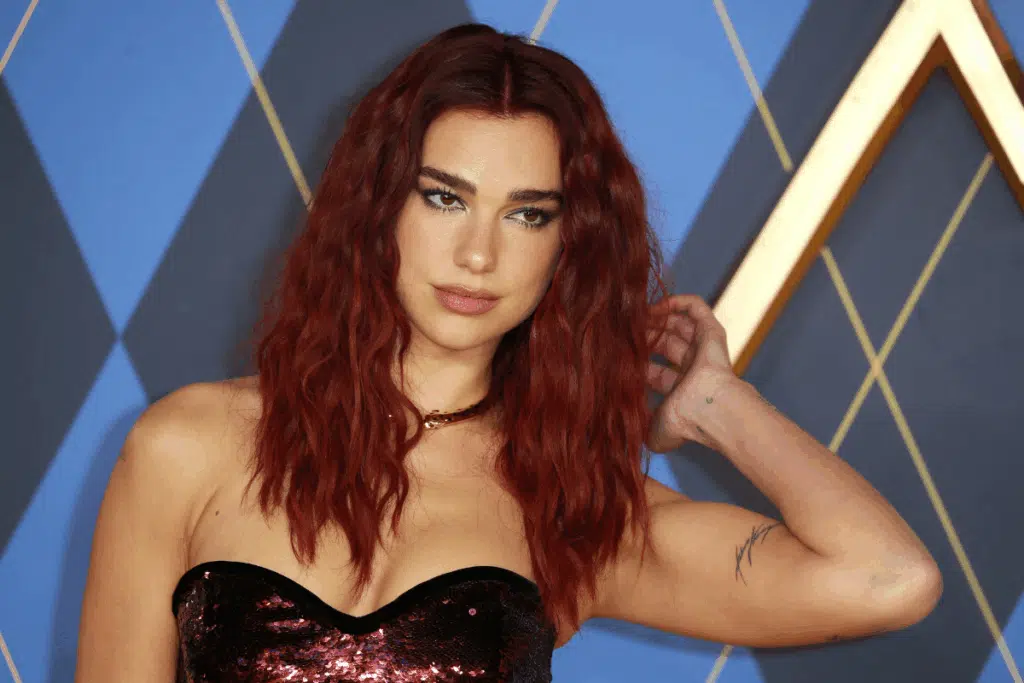 Dua Lipa com cabelo vermelho ondulado e vestido tomara que caia de lantejoulas escuras, ajustando o cabelo com uma das mãos diante de um fundo com padrão geométrico azul e dourado.