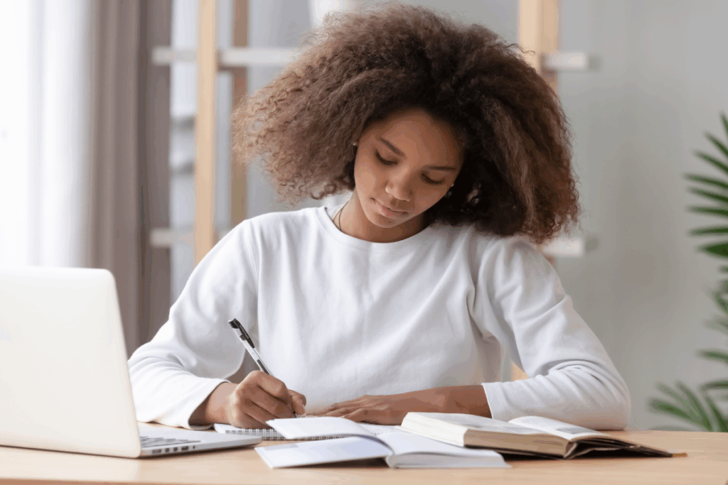 Uma jovem negra está sentada à mesa estudando, segurando uma caneta e escrevendo em um caderno. À sua frente estão livros abertos e, à esquerda, um notebook.