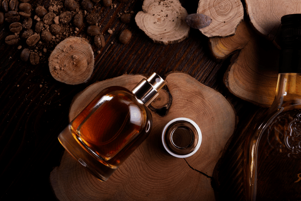Frasco de perfume transparente com líquido âmbar disposto sobre pedaços de madeira, ao lado de grãos de café.