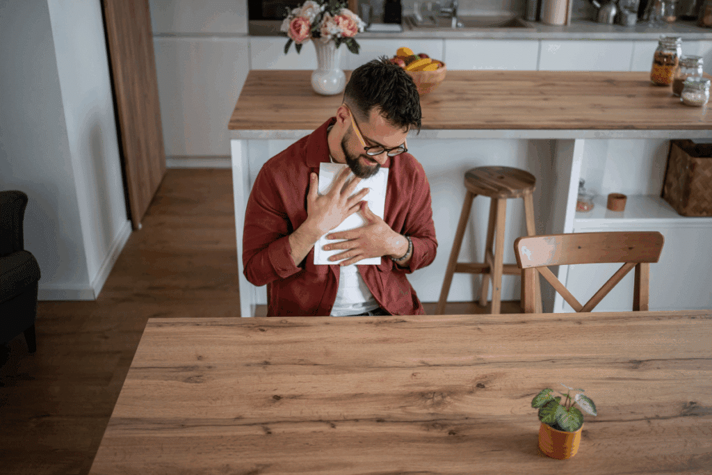 Um homem com barba e óculos, vestindo uma camisa vermelha, está sentado a uma mesa de madeira em uma cozinha moderna. Ele segura um papel branco contra o peito e sorri enquanto olha para baixo. No fundo, há uma bancada com flores, frutas e potes de vidro. Um pequeno vaso de planta está no canto inferior direito da mesa.