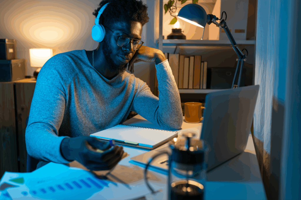 Homem negro usando fones de ouvido brancos e óculos, sentado diante de um notebook em uma mesa de trabalho iluminada por luminária. Ele apoia a cabeça na mão enquanto analisa cadernos, papéis e gráficos em um ambiente com iluminação noturna azulada e acolhedora.