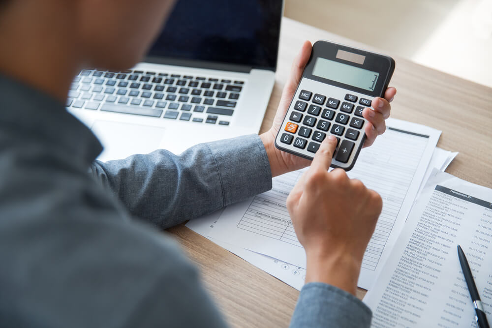 Homem fazendo cálculo em calculadora ao lado de notebook e papéis impressos