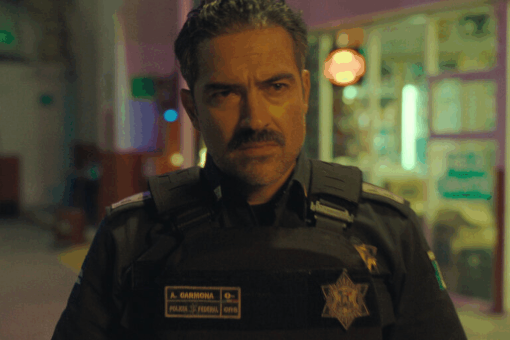 Um close de um homem com bigode vestindo um colete tático de polícia com a inscrição "A. CARMONA" e "POLICIA FEDERAL". Ele tem uma expressão séria e está olhando diretamente para a câmera. O fundo está desfocado, sugerindo um ambiente urbano à noite.