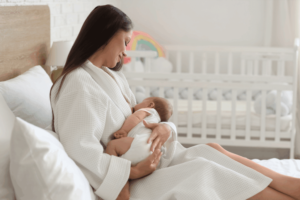 Mulher de ropão branco sentada na cama amamentando seu bebê recém-nascido no colo. Ao fundo, vê-se um berço infantil e decoração em tons claros.