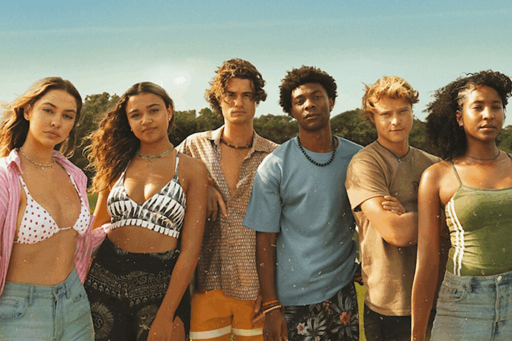 Um grupo de seis jovens está de pé lado a lado ao ar livre, olhando para a câmera. Eles estão vestidos com roupas de verão casuais, como biquínis, camisas de botão abertas e camisetas. O fundo é um campo gramado com árvores e um céu claro.