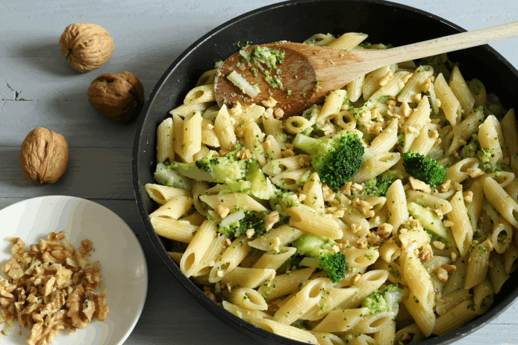 Frigideira com macarrão penne misturado a pedaços de brócolis e nozes picadas, acompanhada de uma colher de pau. Ao lado, sobre uma mesa de madeira, há nozes inteiras e um pratinho com nozes trituradas.