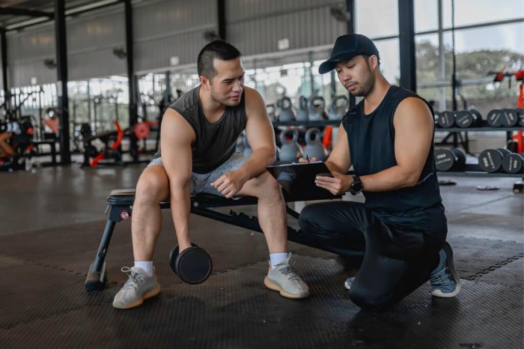 Fotografia de dois homens em uma academia. À esquerda, um homem vestindo regata cinza-escuro e calção claro está sentado em um banco de musculação, segurando um halter com uma das mãos. À direita, um personal trainer ajoelhado, usando boné e regata pretos, segura uma prancheta enquanto orienta o aluno. Ao fundo, veem-se anilhas e outros equipamentos da academia.
