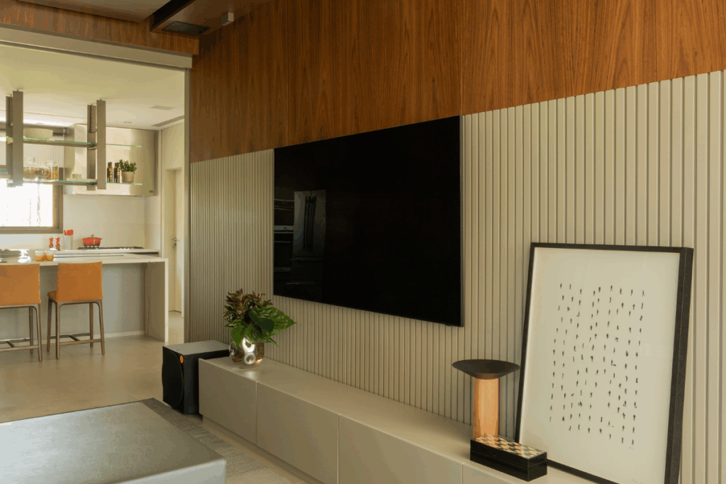 Painel de TV em sala integrada, combinando acabamento em madeira no topo e revestimento canelado claro embaixo, com rack cinza, vaso de planta e um quadro apoiado no chão.