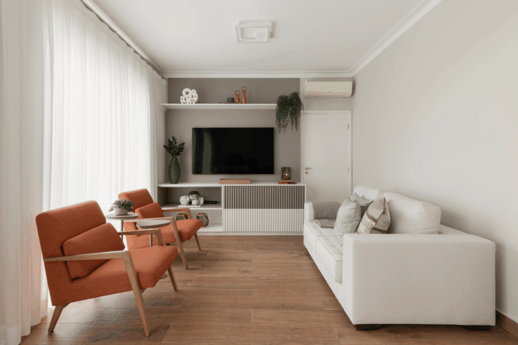 Foto de uma sala de estar iluminada com piso de madeira, sofá branco à direita, duas poltronas laranjas à esquerda e painel de TV ao fundo.
