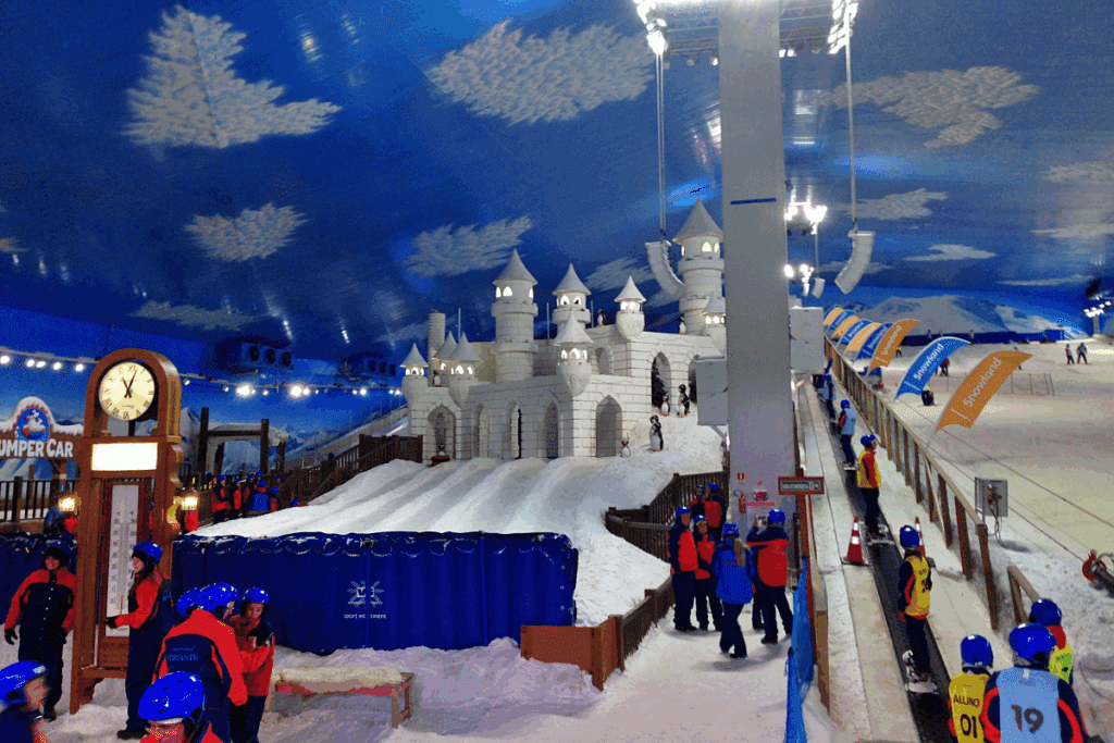 Vista interna do parque temático coberto Snowland, mostrando pessoas com roupas de inverno e capacetes azuis, uma pista de esqui, uma esteira rolante e um grande castelo de gelo ao centro.