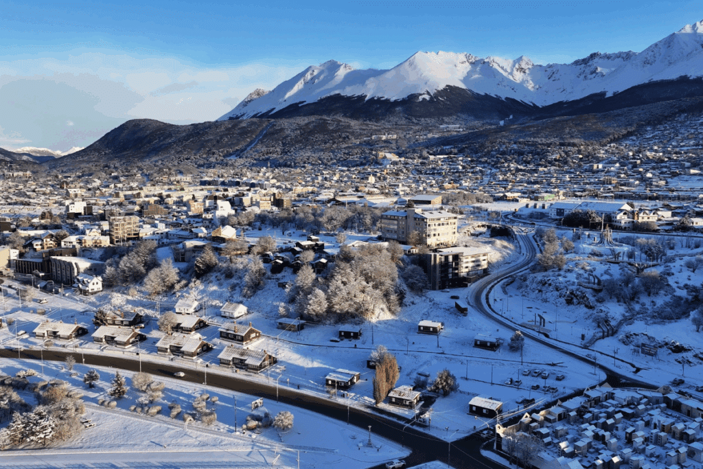 Vista aérea da cidade de Ushuaia coberta de neve, com ruas, casas e edifícios em primeiro plano e grandes montanhas nevadas ao fundo sob um céu claro.