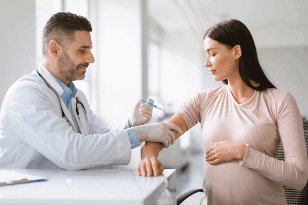 Médico branco de jaleco branco aplicando uma vacina no braço de uma mulher grávida, que está sentada e com a mão apoiada na barriga.