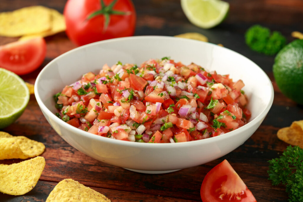 Tomate, cebola-roxa e cheiro-verde em recipiente branco fundo ao lado de ingredientes frescos como tomate e limão