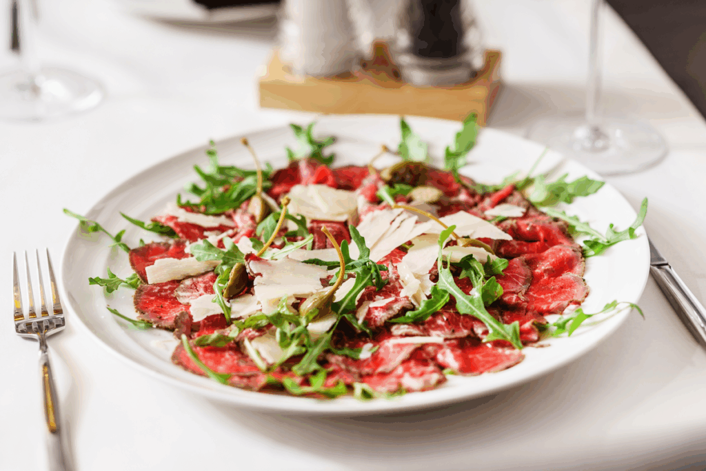 Prato branco com carpaccio de carne bovina coberto por lascas de parmesão, folhas de rúcula e alcaparras, servido em mesa posta com toalha branca.