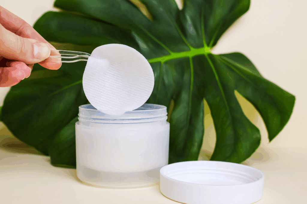 Mão segurando pinça e pegando toner pads de pote branco. folha verde de planta ao fundo