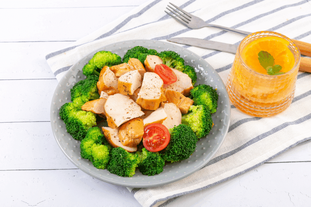 Prato com floretes de brócolis, frango temperado com pimenta-do-reino e tomate, servido sobre uma mesa de madeira. Ao lado, um copo com suco amarelo