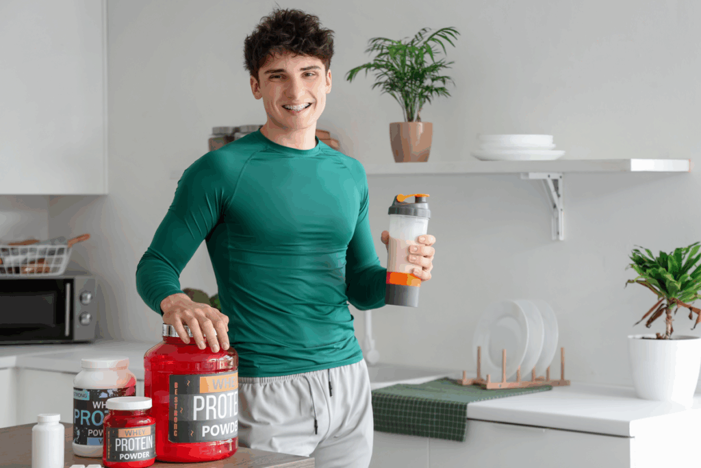 Jovem de camiseta verde sorrindo na cozinha com uma coqueteleira de shake na mão, apoiado em potes grandes de whey protein.