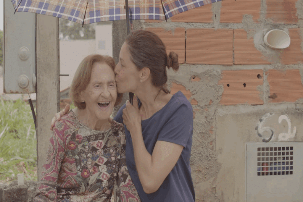 Mulher idosa usando camisa estampada sorrindo e uma mulher mais jovem segurando um guarda-chuva estampado e com cabelo preso em coque e usando camiseta azul beijando o rosto da mulher idosa