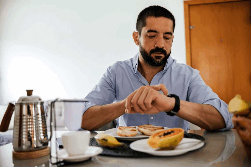 Homem com cabelo curto, barba, usando camisa social azul, comendo e olhando hora no relógio