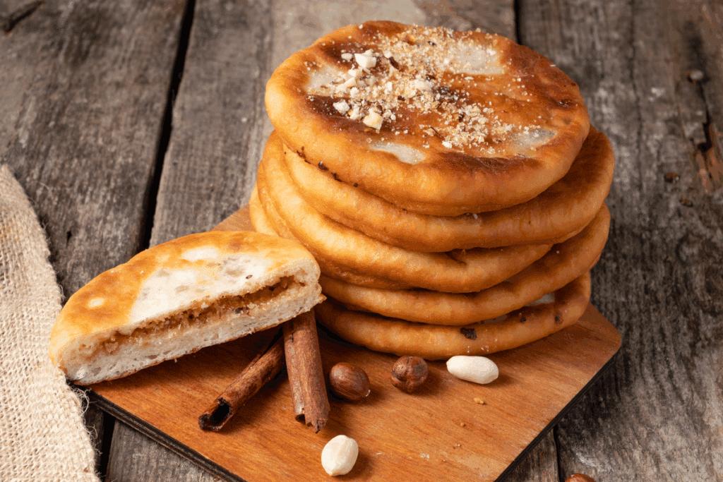 Pilha de hottoks dourados sobre uma tábua de madeira, acompanhada de um hottok partido ao meio que revela o recheio de açúcar, canela e castanhas, com paus de canela e amendoins ao lado