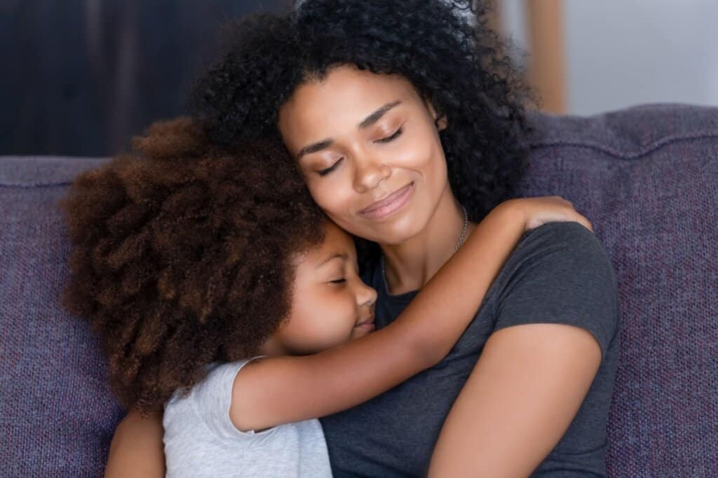 mãe e filha pequena de camiseta cinza, de olhos fechados e abraçadas. elas estão sentadas em sofá cinza