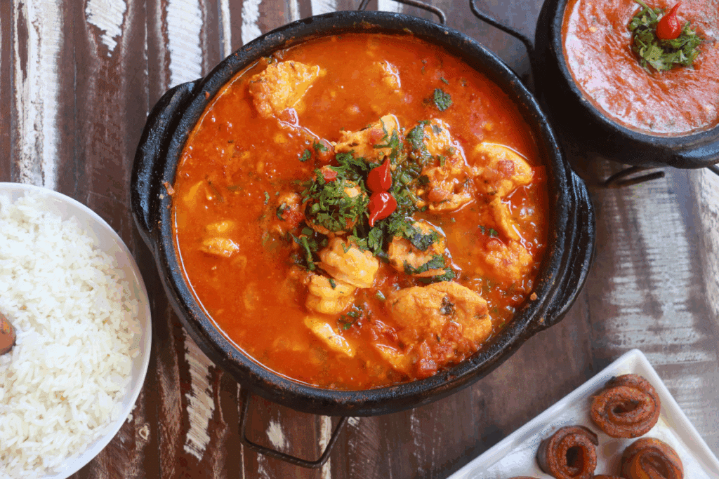 A imagem mostra uma moqueca de peixe servida em uma panela de barro preta, com caldo de coloração avermelhada e pedaços generosos de peixe. O prato está finalizado com ervas frescas picadas e pequenas pimentas vermelhas, que dão destaque ao centro da preparação.