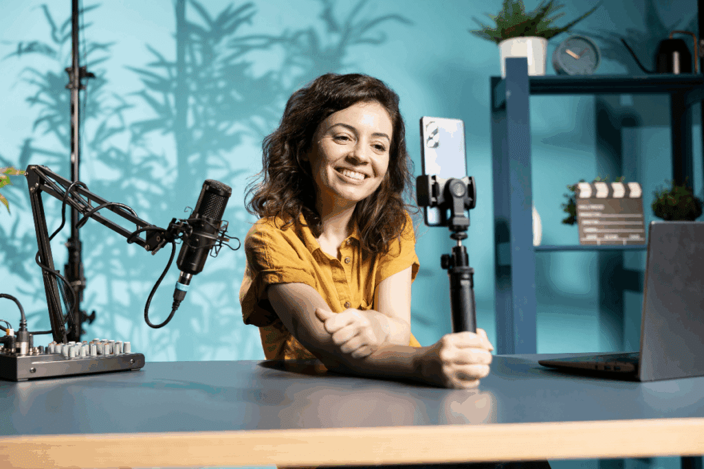 A imagem mostra uma mulher sorridente gravando conteúdo com um celular, posicionado em um suporte sobre a mesa. Ao lado dela, há um microfone profissional articulado, uma mesa de áudio e um notebook, formando uma estrutura voltada para produção audiovisual.