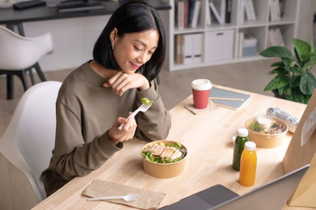 A imagem mostra uma mulher sentada à mesa enquanto come uma salada em um ambiente moderno e aconchegante. Sobre a mesa há refeições leves, bebidas naturais e um notebook