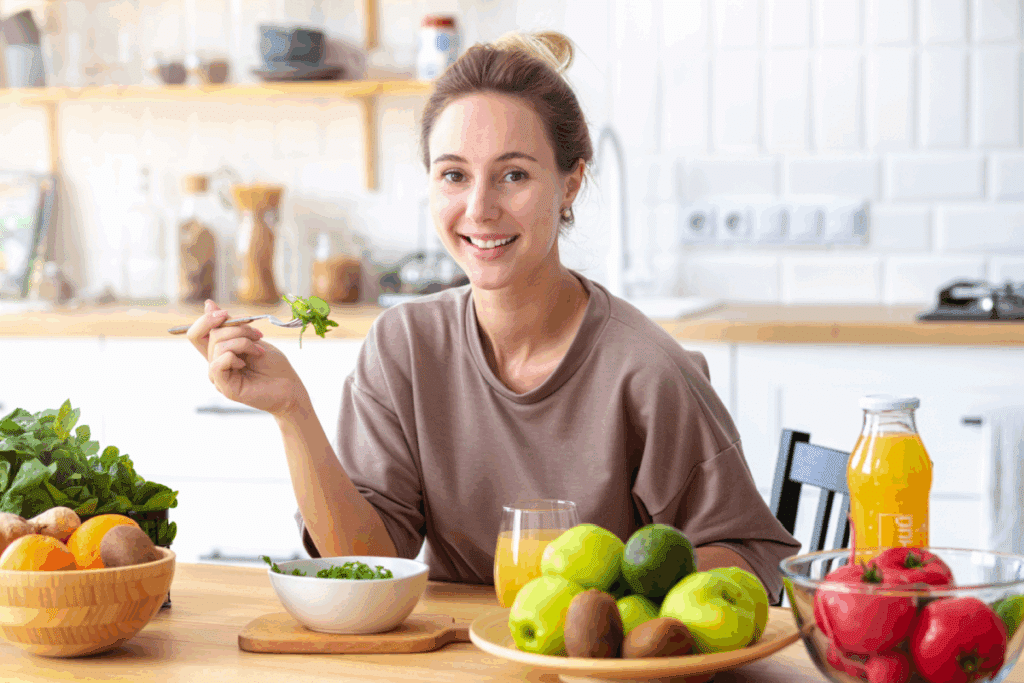 A imagem mostra uma mulher sentada à mesa em uma cozinha clara e moderna, sorrindo enquanto segura um garfo com folhas de salada prestes a serem consumidas. Sobre a mesa há uma tigela de salada, frutas como maçãs, laranjas e kiwis, folhas verdes frescas, tomates e uma garrafa de suco natural, compondo um cenário de alimentação saudável.