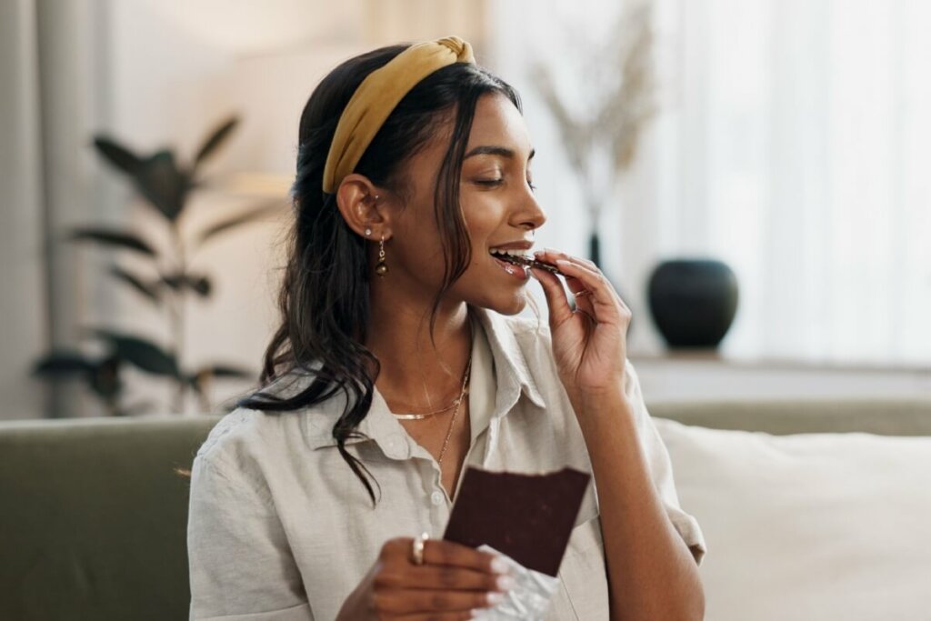 Mulher sorridente com tiara amarela no cabelo comendo um pedaço de chocolate de barra enquanto segura a embalagem