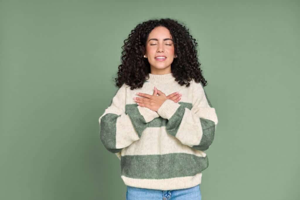 A imagem mostra uma mulher com os olhos fechados, mãos cruzadas sobre o peito, em um gesto que transmite calma e introspecção. Ela veste um suéter em tons claros e está diante de um fundo verde suave, que reforça a sensação de tranquilidade.