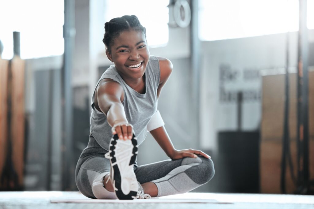 A imagem mostra uma mulher usando roupas esportivas enquanto realiza um alongamento em uma academia. Sentada sobre um colchonete, ela estende uma das pernas à frente e segura o pé com a mão, executando um exercício de flexibilidade, enquanto sorri
