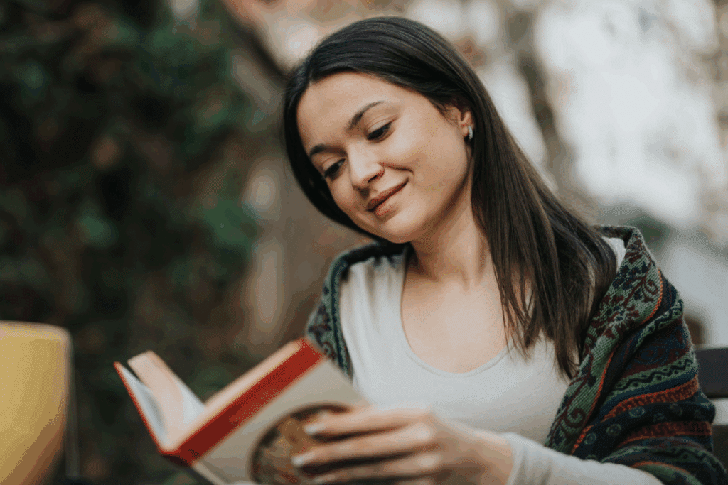 Mulher com cabelo liso, solto usando camisa de manga longa cinza e com coberta nos ombros lendo livro em local aberto